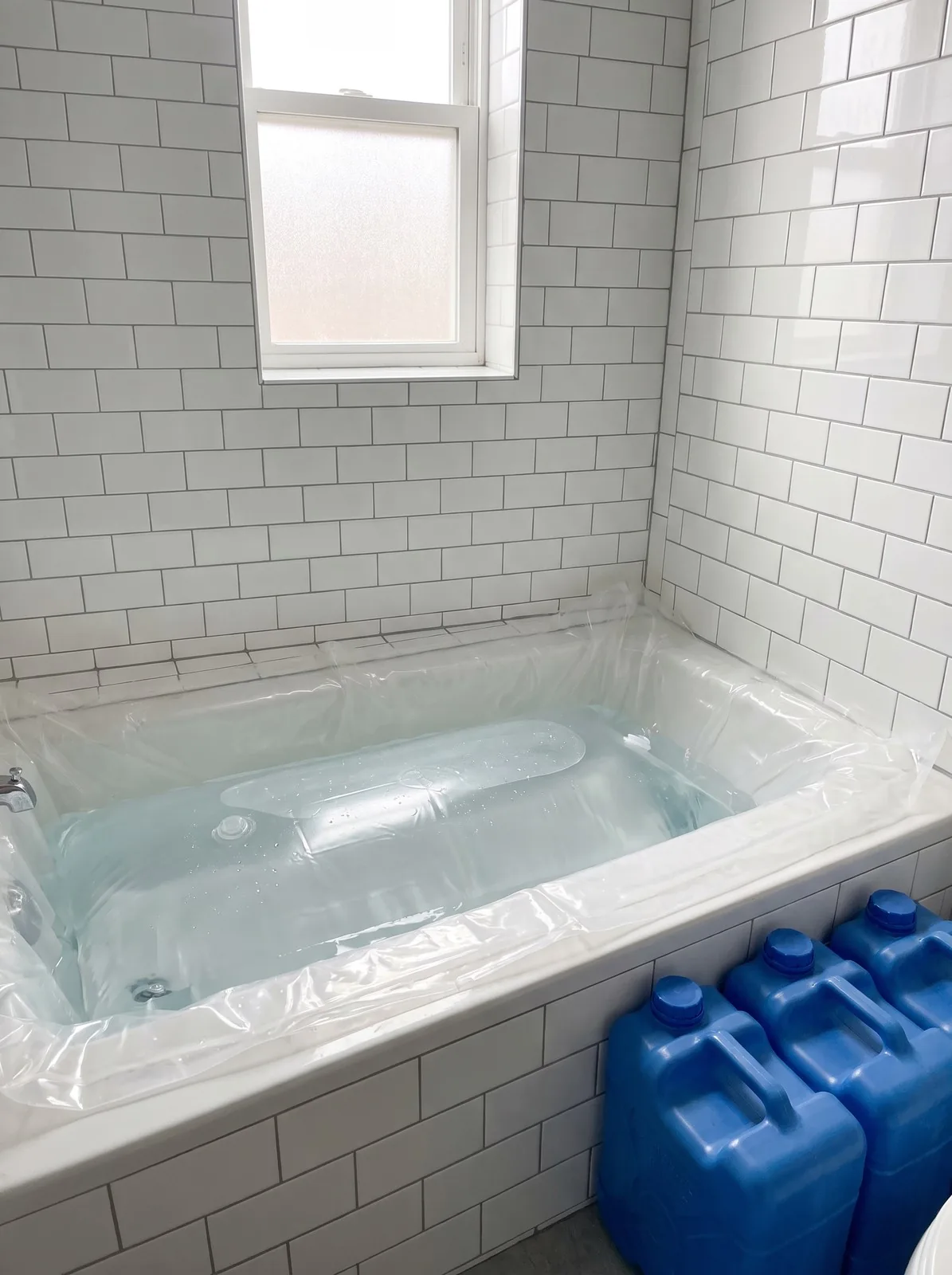 A bathtub lined with a clear water-storage bladder full of water beside stacked blue water jugs in a tiled bathroom