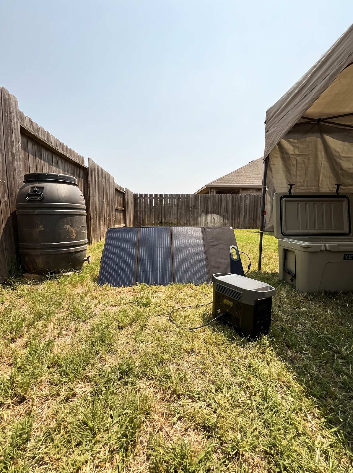 A folding solar panel in a backyard charging a portable power station beside a rain barrel and a cooler