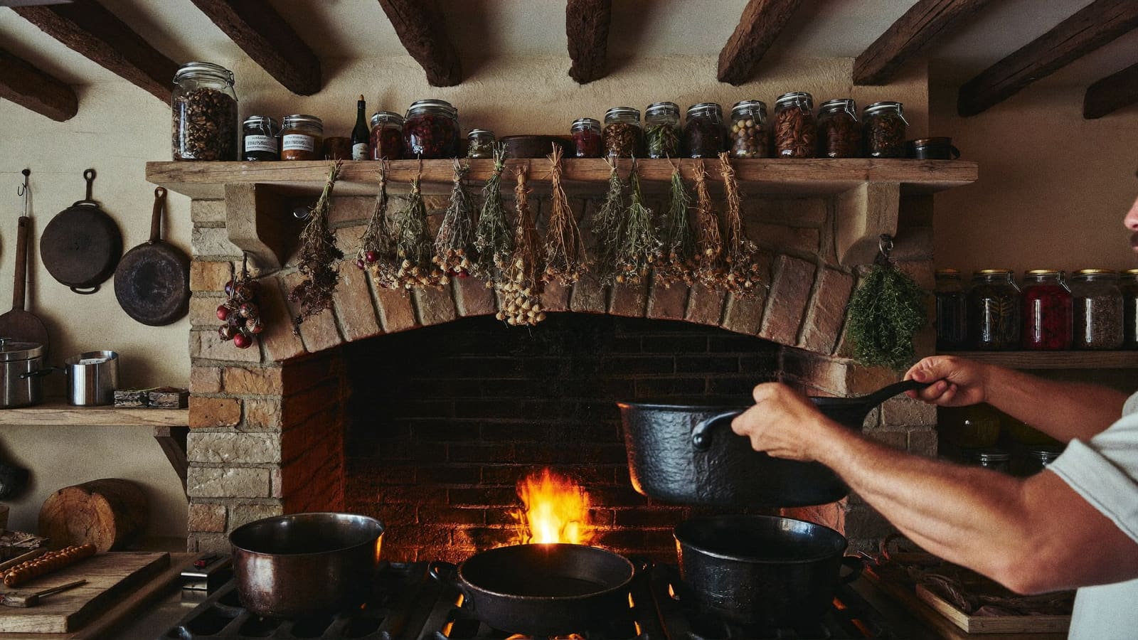 Homestead kitchen cooking with cast iron over open fireplace, dried herbs hanging above, rustic frontier aesthetic